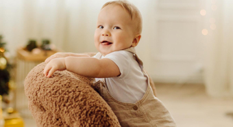 Toddler smiles while standing on a cozy looking fuzzy brown colored chair in a house setting. 