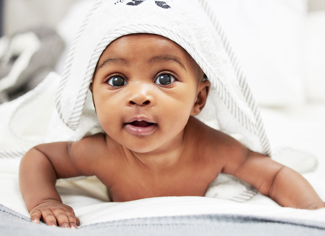 A baby with brown skin and brown eyes lying on its stomach holding its head up smiling at the camera with a bath towel lying over its head and body.