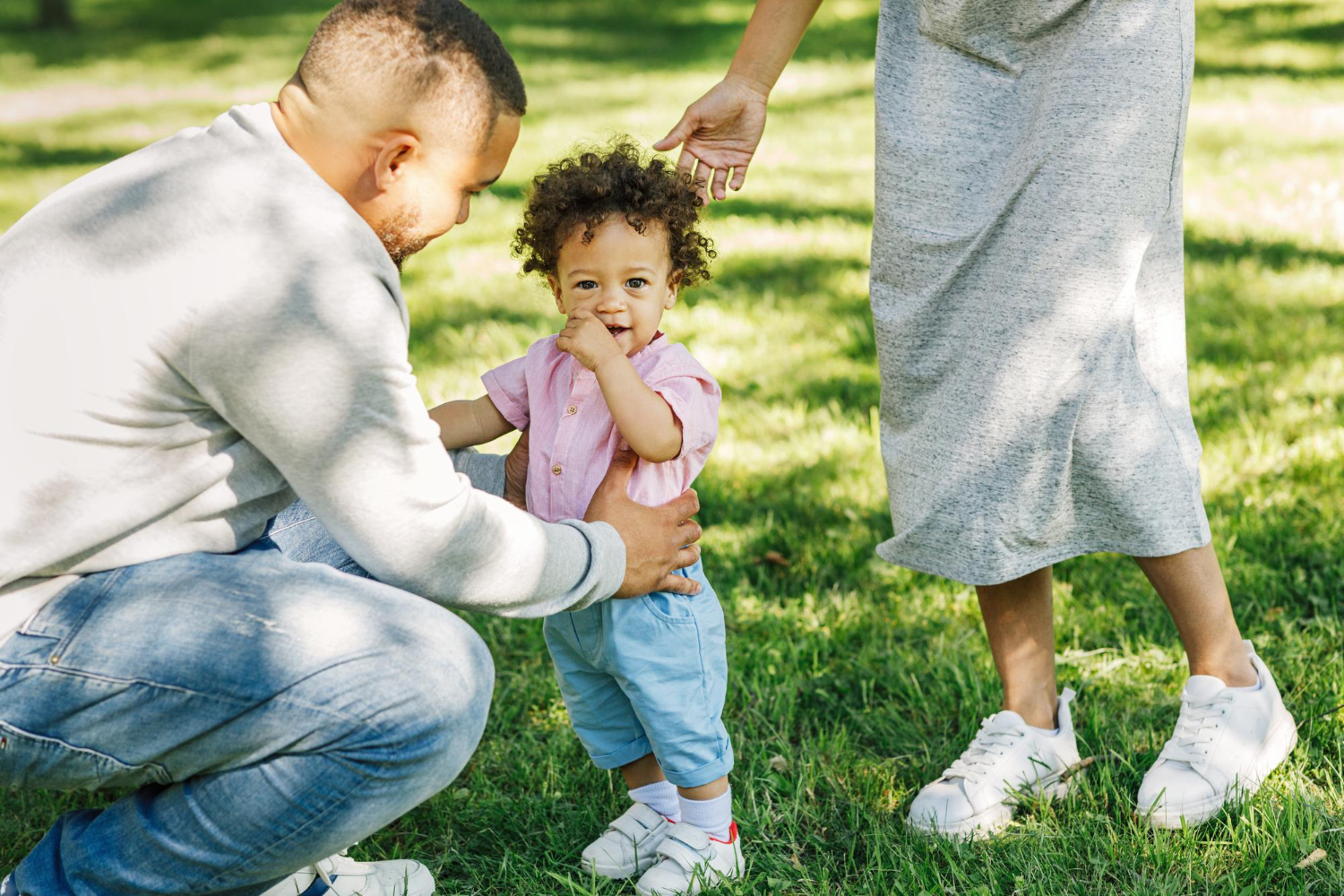 Toddler with curly hair stands with two adults in a sunny park setting. 