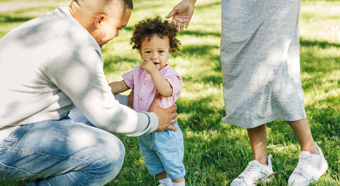 Toddler with curly hair stands with two adults in a sunny park setting. 