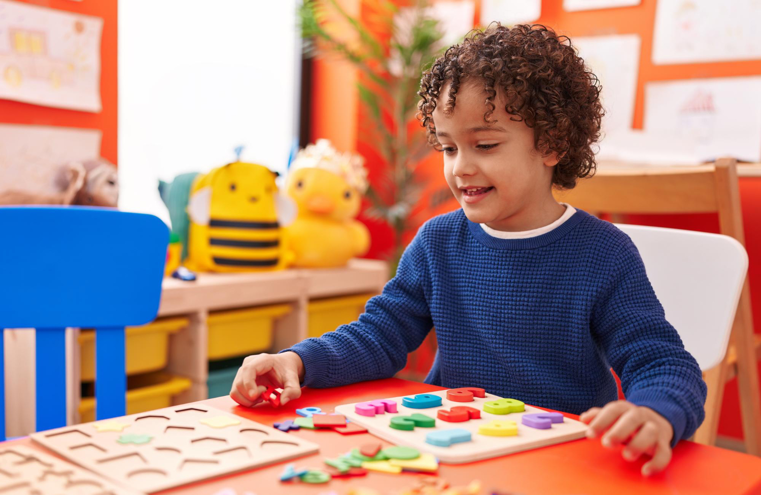 Young boy sitting in colorful classroom setting, wearing blue long sleeved shirt, smiles while playing with blocks. 