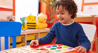 Young boy sitting in colorful classroom setting, wearing blue long sleeved shirt, smiles while playing with blocks. 