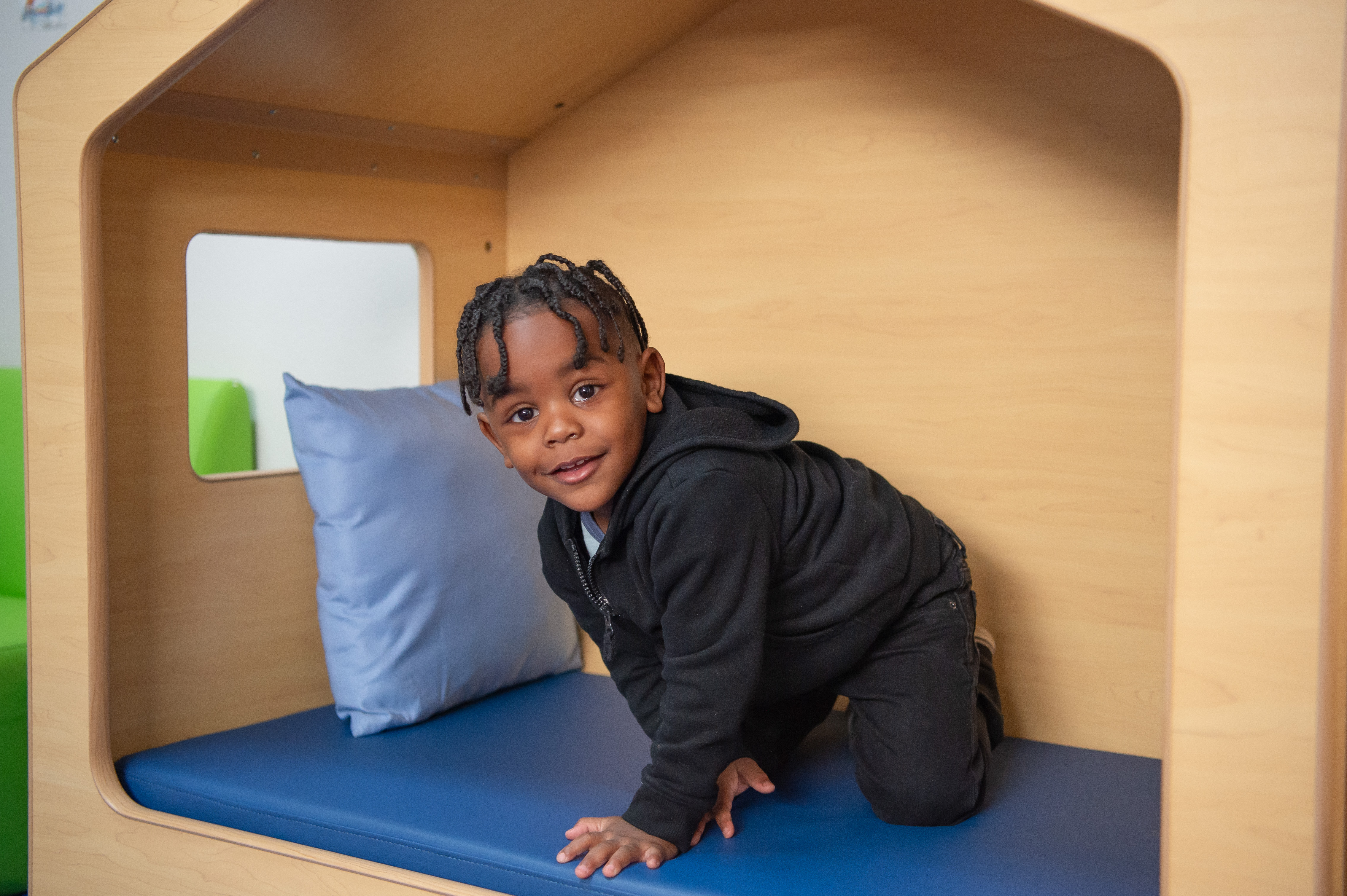 Young boy, in an indoor play house with blue cushions, smiles at the camera. Wearing all black clothing. 