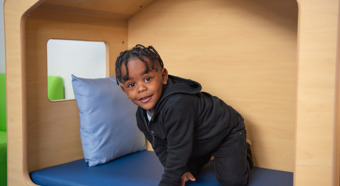 Young boy, in an indoor play house with blue cushions, smiles at the camera. Wearing all black clothing. 