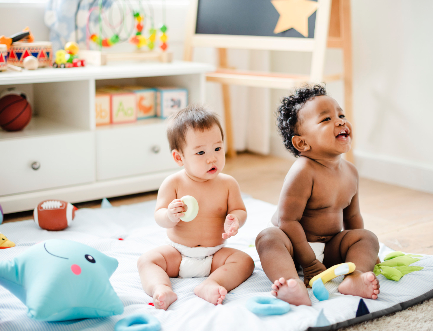 Two toddlers sitting in diapers on a blanket on the floor, smiling, in a comfy at home setting. 