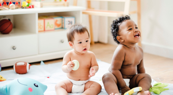 Two toddlers sitting in diapers on a blanket on the floor, smiling, in a comfy at home setting. 