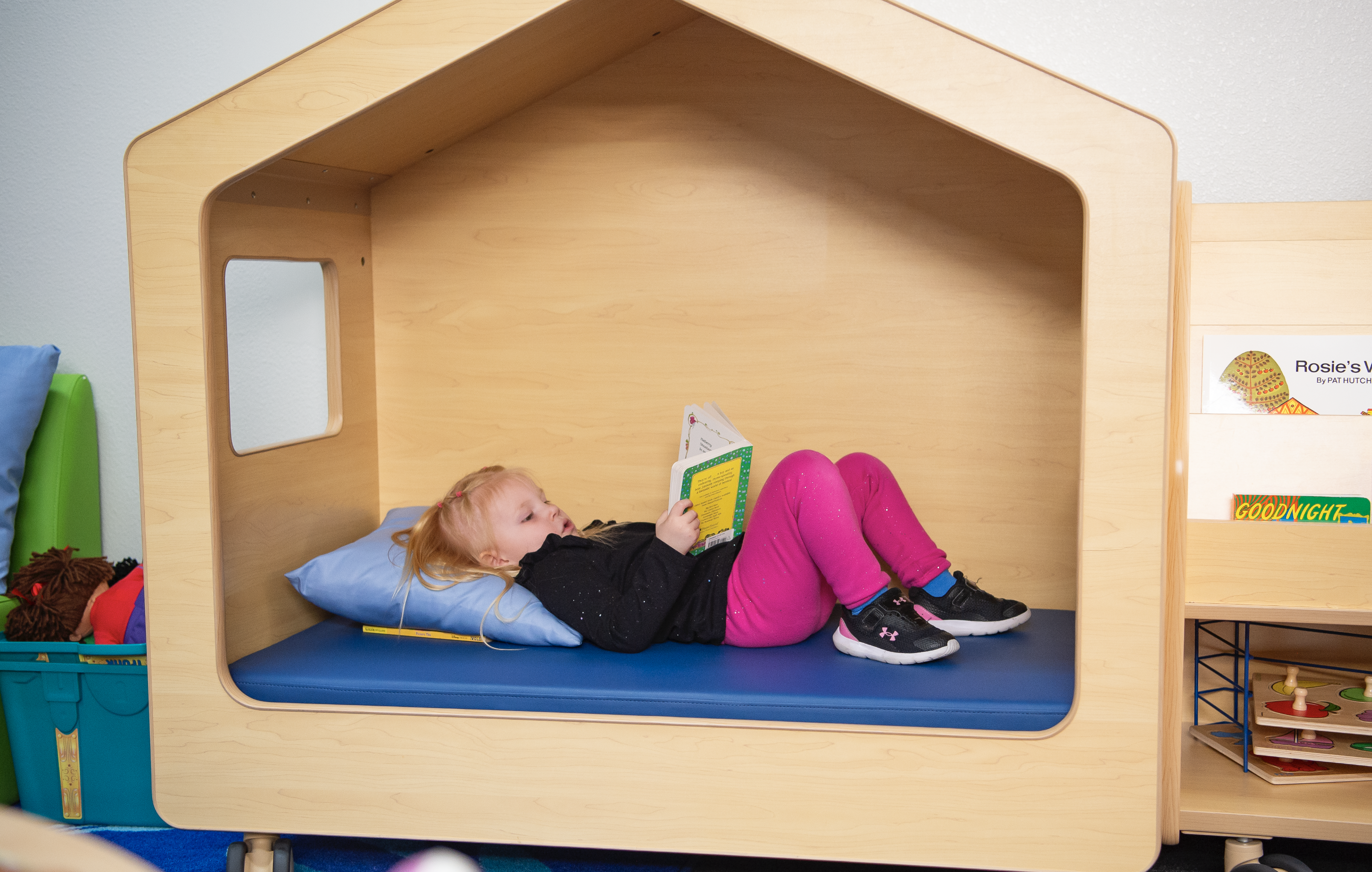 Little girl laying In a playhouse bed while reading a book. Wearing pink pants.