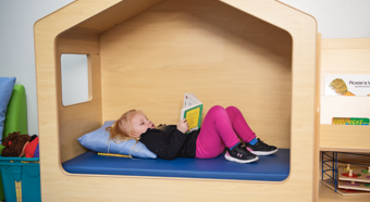 Little girl laying In a playhouse bed while reading a book. Wearing pink pants.