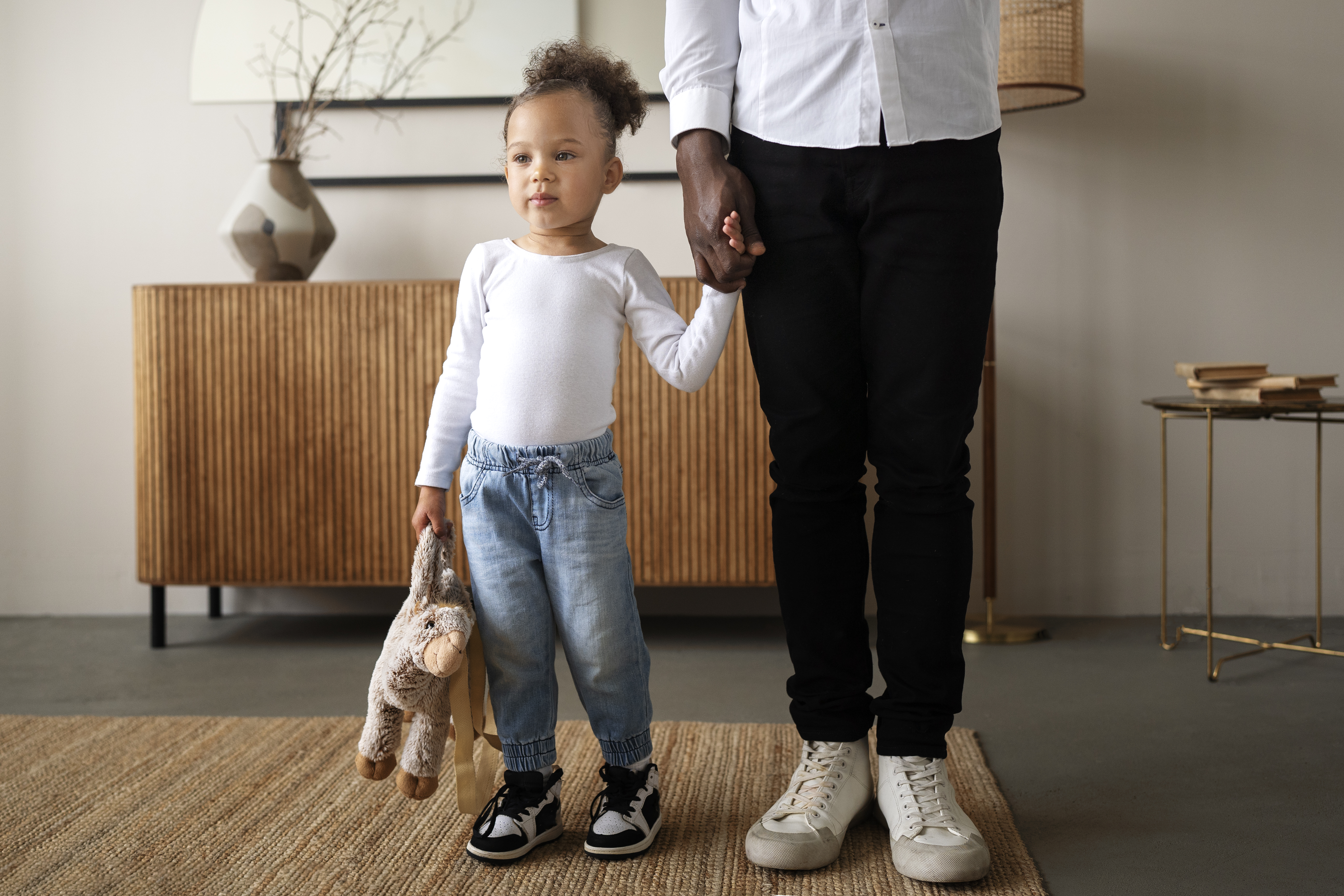 Little girl wearing white long sleeve shirt, jeans, holding a stuffed animal holds the hand of an adult figure. Indoor house setting.