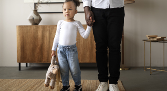 Little girl wearing white long sleeve shirt, jeans, holding a stuffed animal holds the hand of an adult figure. Indoor house setting.