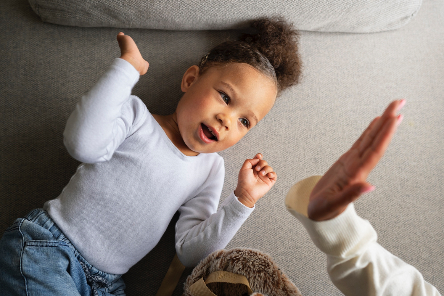 Happy toddler lays on floor about to high five an adult, smiling.