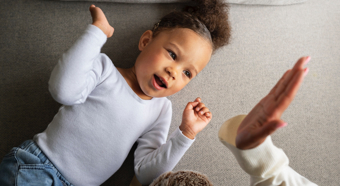 Happy toddler lays on floor about to high five an adult, smiling.