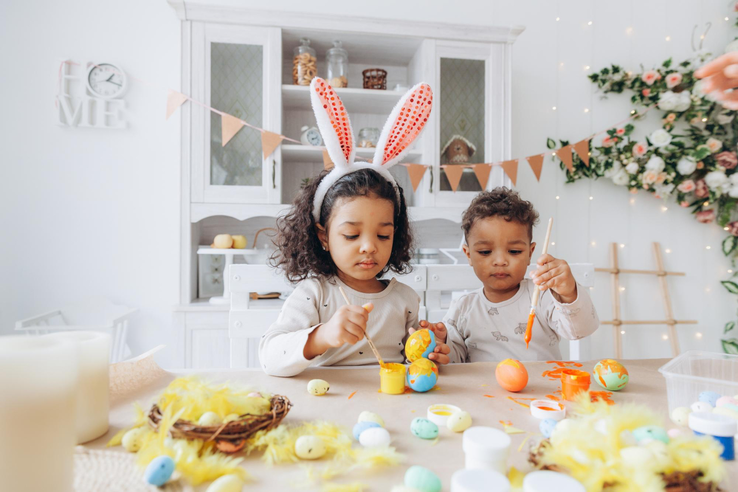 Two little kids sitting at a table in a home setting, painting easter eggs and one is wearing easter bunny ears.