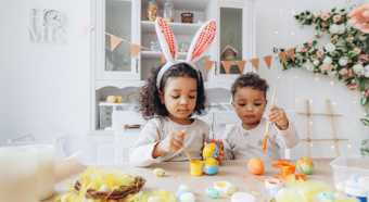 Two little kids sitting at a table in a home setting, painting easter eggs and one is wearing easter bunny ears.