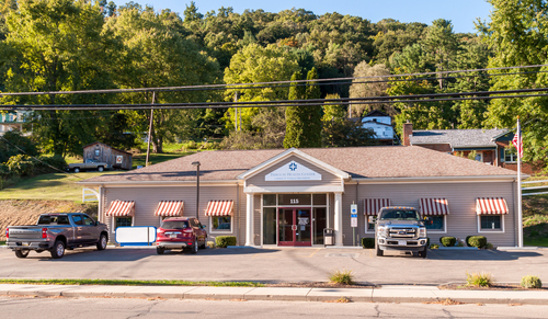 Exterior shot of rural building, green trees in background, 3 cars parked in front of the single story building. 