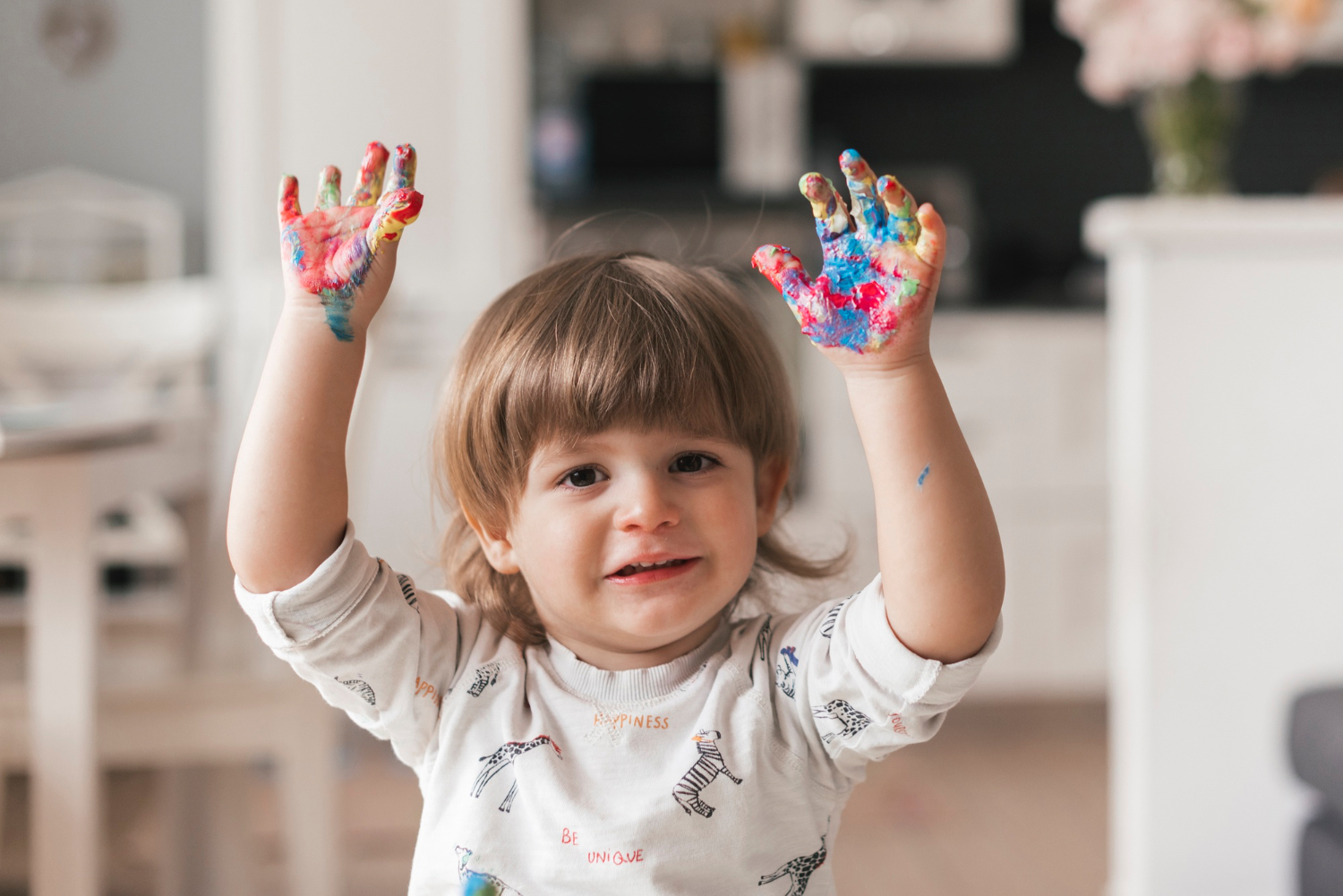 Toddler with rainbow paint on his hands smiles and holds his hands in the air. 