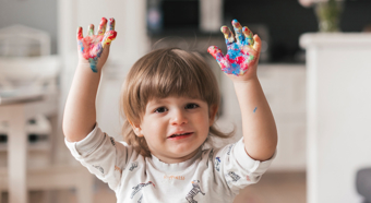 Toddler with rainbow paint on his hands smiles and holds his hands in the air. 