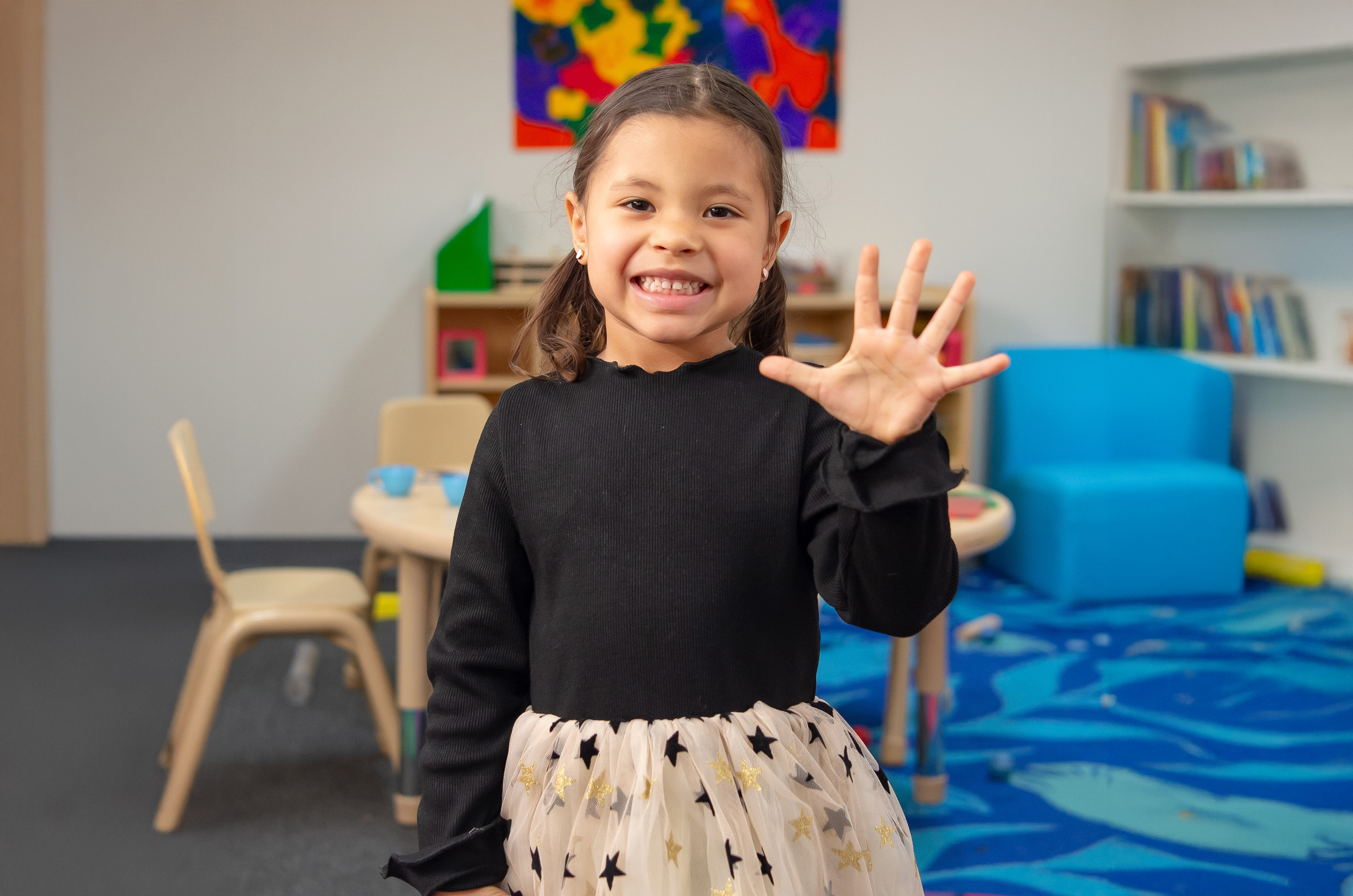 Young girl smiles at camera and hold up hand with five fingers up, in a colorful classroom setting