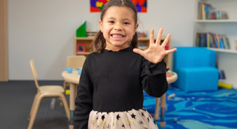 Young girl smiles at camera and hold up hand with five fingers up, in a colorful classroom setting