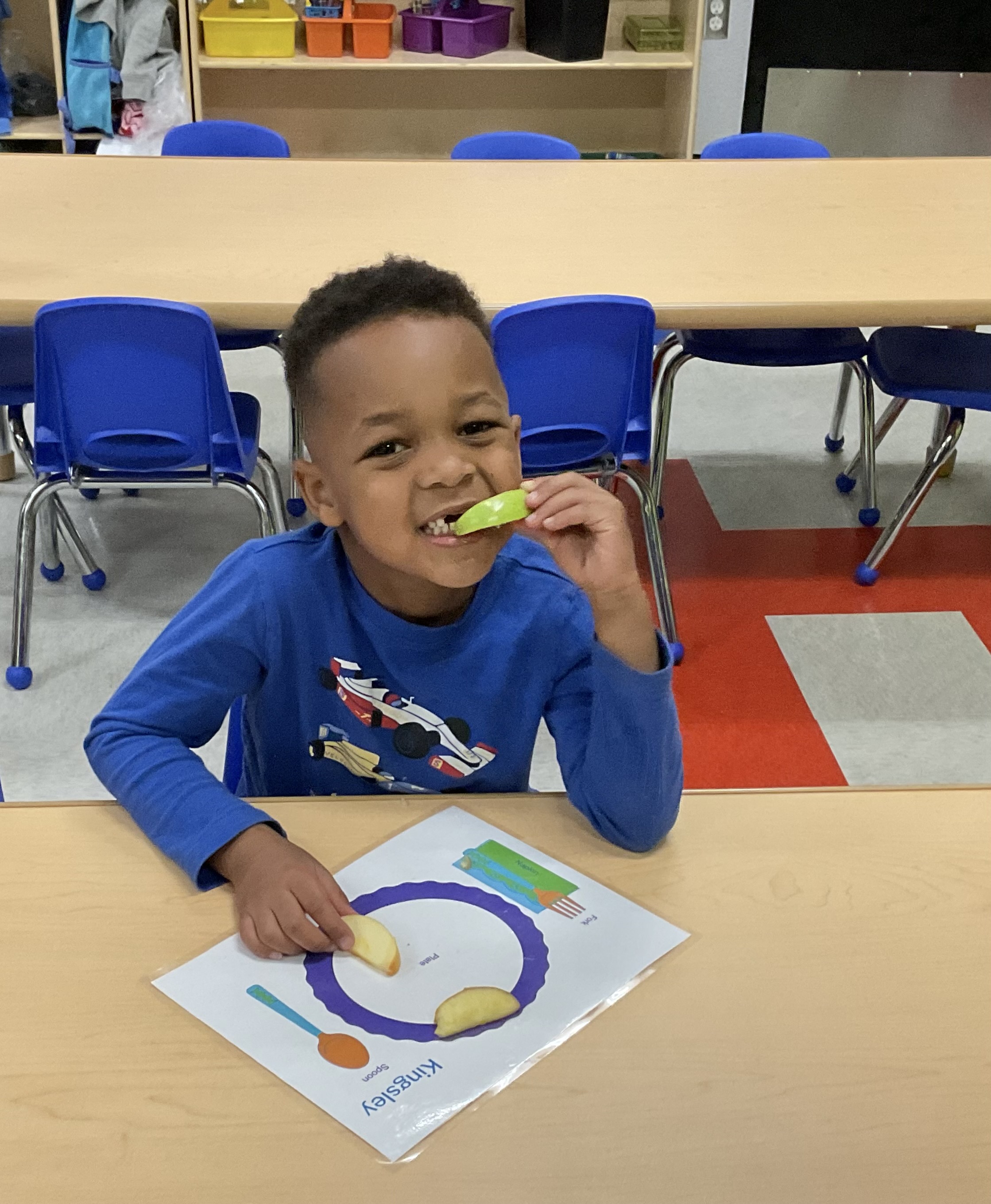 Toddler boy sitting down eating an apple slice smiles at camera. 