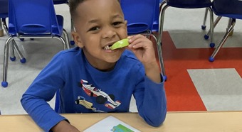 Toddler boy sitting down eating an apple slice smiles at camera. 