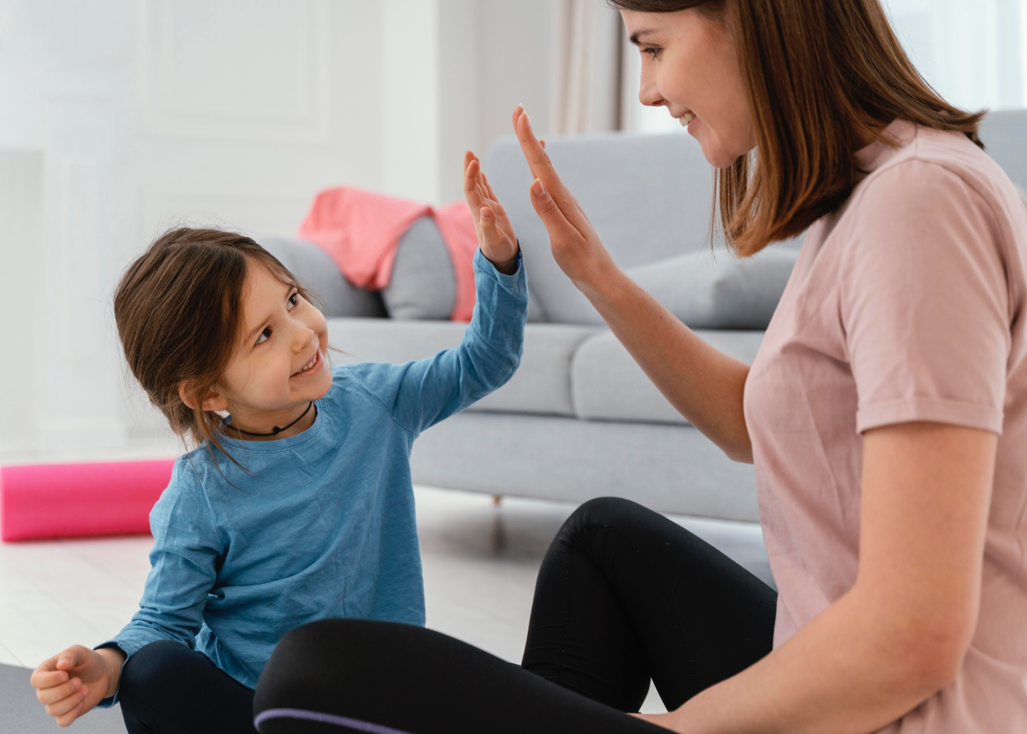 Little girl in blue shirt high fives adult women in pink shirt, both sitting on the floor smiling at each other. 
