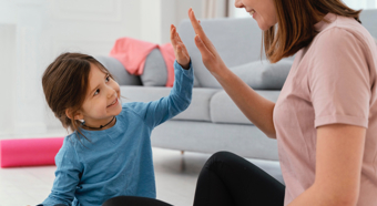 Little girl in blue shirt high fives adult women in pink shirt, both sitting on the floor smiling at each other. 