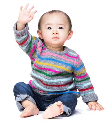 A baby with light brown skin and hair, sitting up waving at the camera, and smiling slightly. She has on a rainbow sweater and jeans.