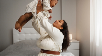 Adult female holding baby in the air, both looking at each other smiling. In a comfy home setting.
