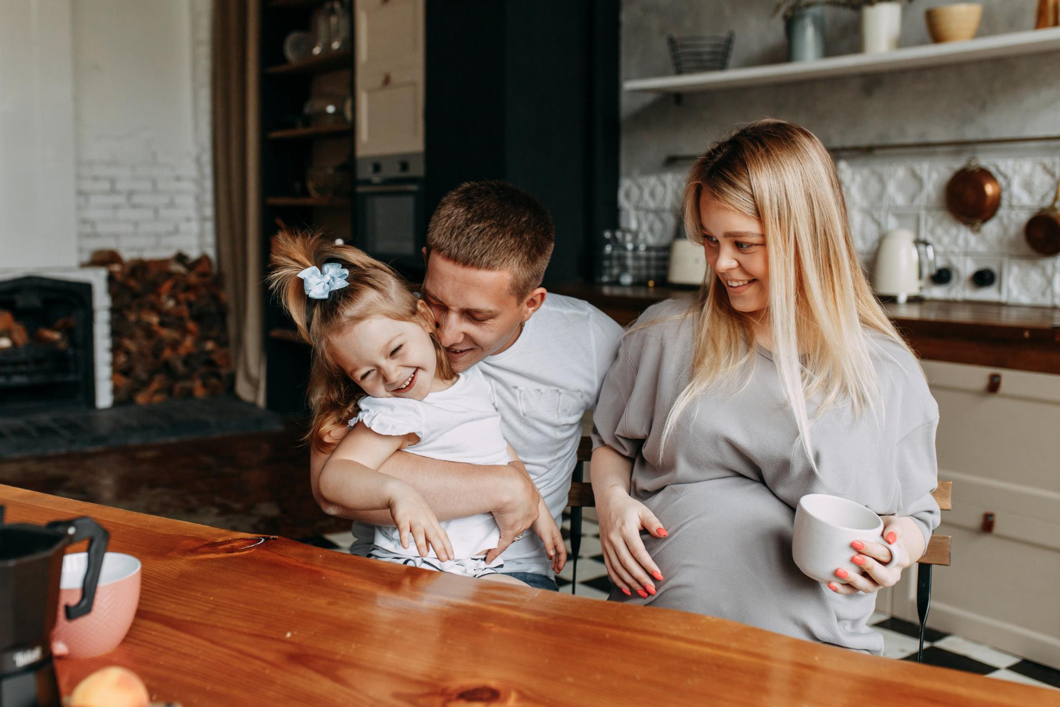 Adult male, female and toddler all sitting at a wood table in a kitchen setting, all laughing and smiling, having coffee.