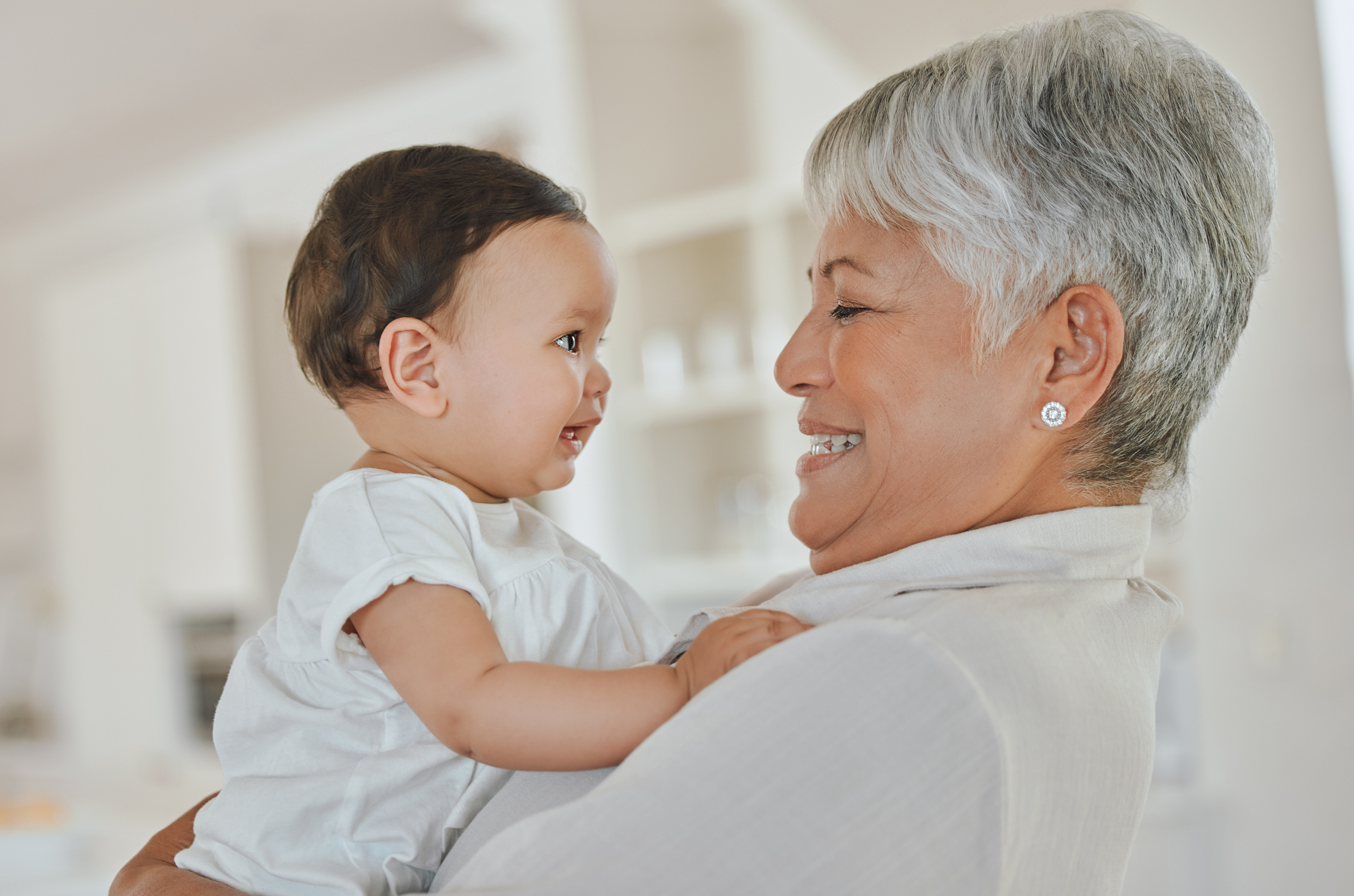 Grandma adult smiles at baby she is holding in her arms. Both are happy and wearing all white, in a bright house setting. 