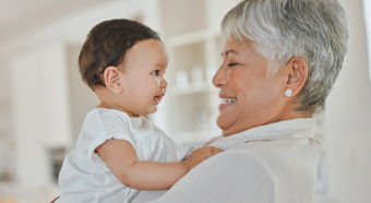 Grandma adult smiles at baby she is holding in her arms. Both are happy and wearing all white, in a bright house setting. 