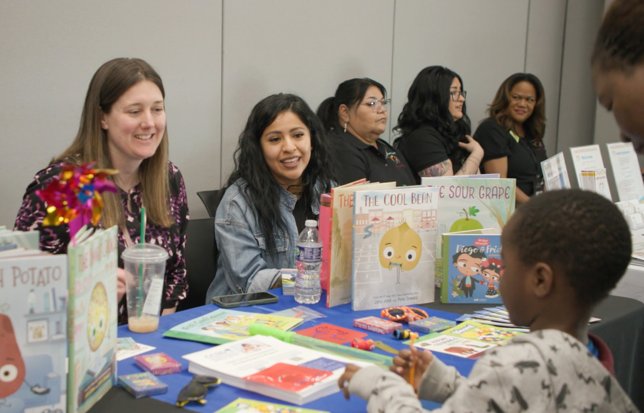 Two women sitting at a booth at a Screening Event smile while talking to young toddler kid at the booth. 