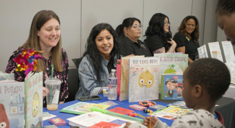Two women sitting at a booth at a Screening Event smile while talking to young toddler kid at the booth. 