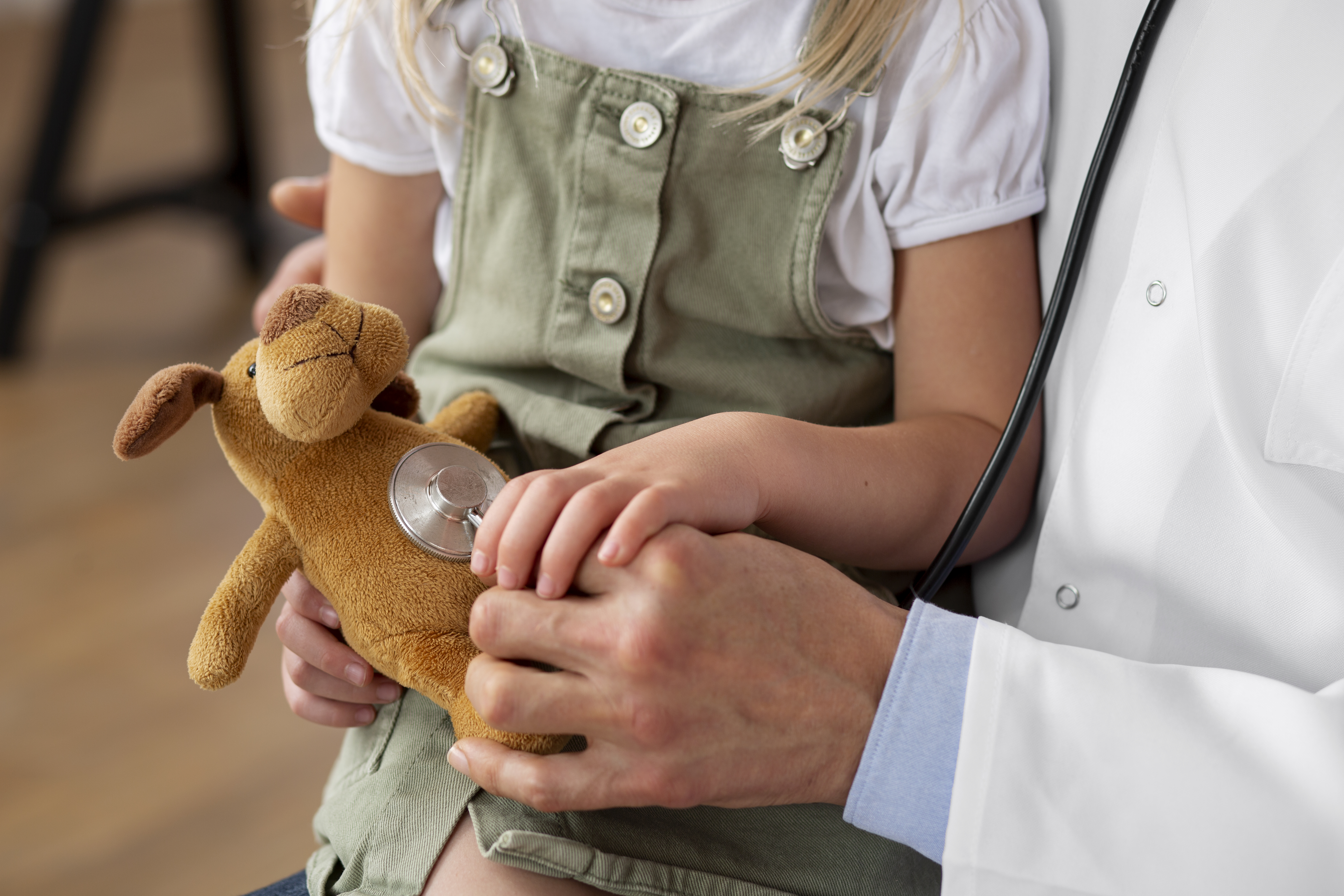 Close up of medical professional in white lab coat with young girl in their lap and holding her hand. Girl is holding a stuffed animal and wearing a dark green outfit. 