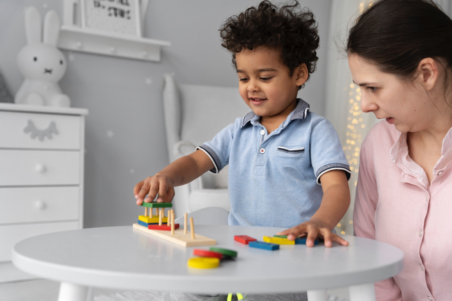 Young boy wearing light blue shirt plays with toys while adult female next to her wearing pink shirt is talking to him. 