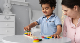 Young boy wearing light blue shirt plays with toys while adult female next to her wearing pink shirt is talking to him. 