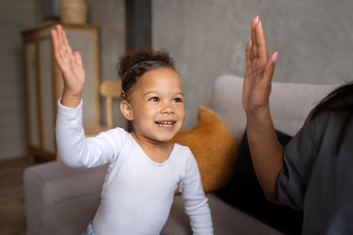 Smiling toddler girl, wearing white long sleeve shirt, hold up her hand ready for a high five from the adult figure to her right. In a comfy home setting. 