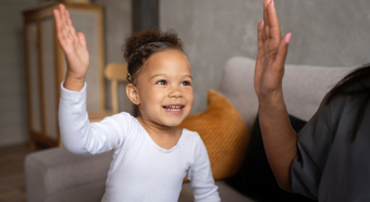 Smiling toddler girl, wearing white long sleeve shirt, hold up her hand ready for a high five from the adult figure to her right. In a comfy home setting. 