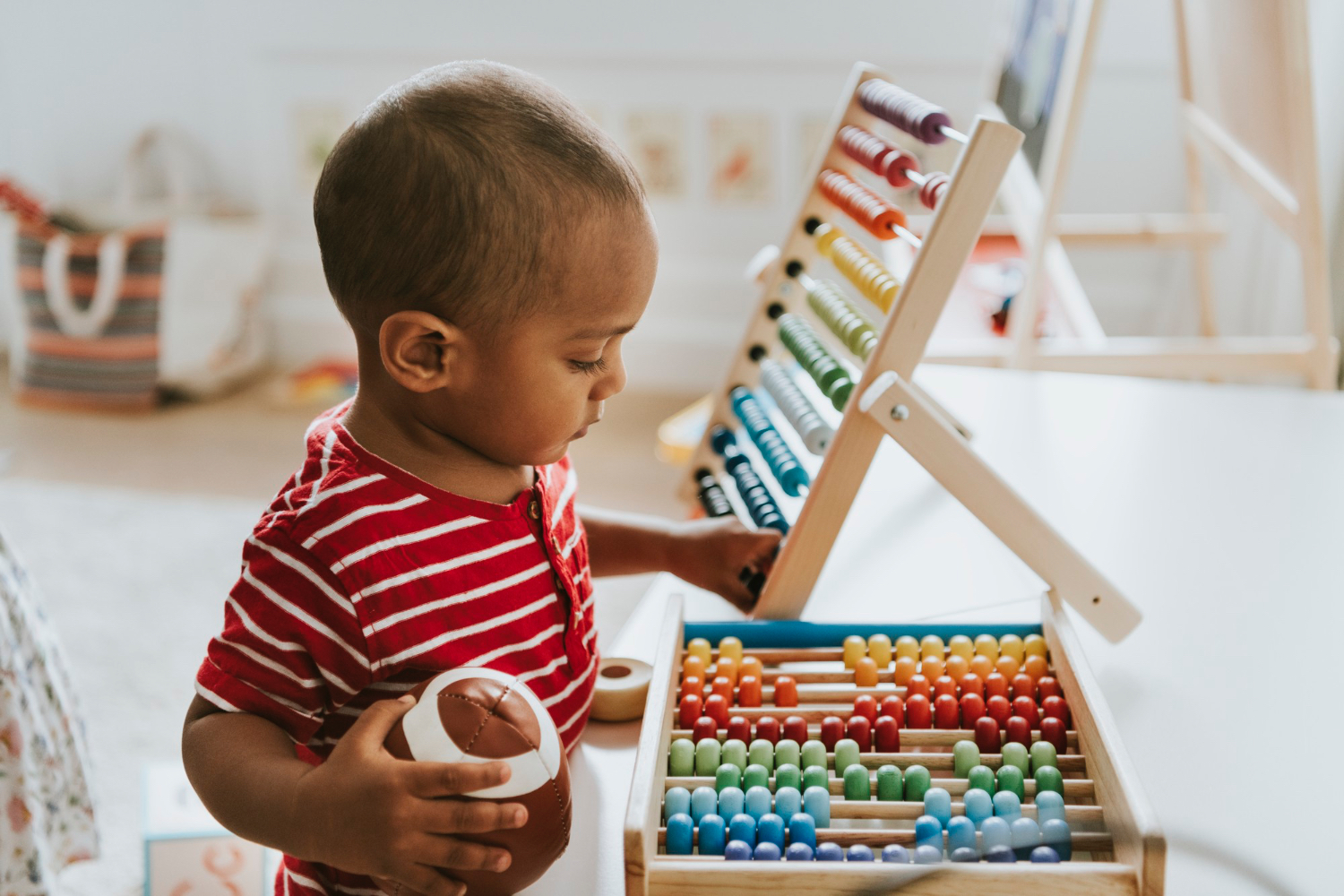 Toddler playing with rainbow children's games, wearing red stripped shirt and holding a toy football. 