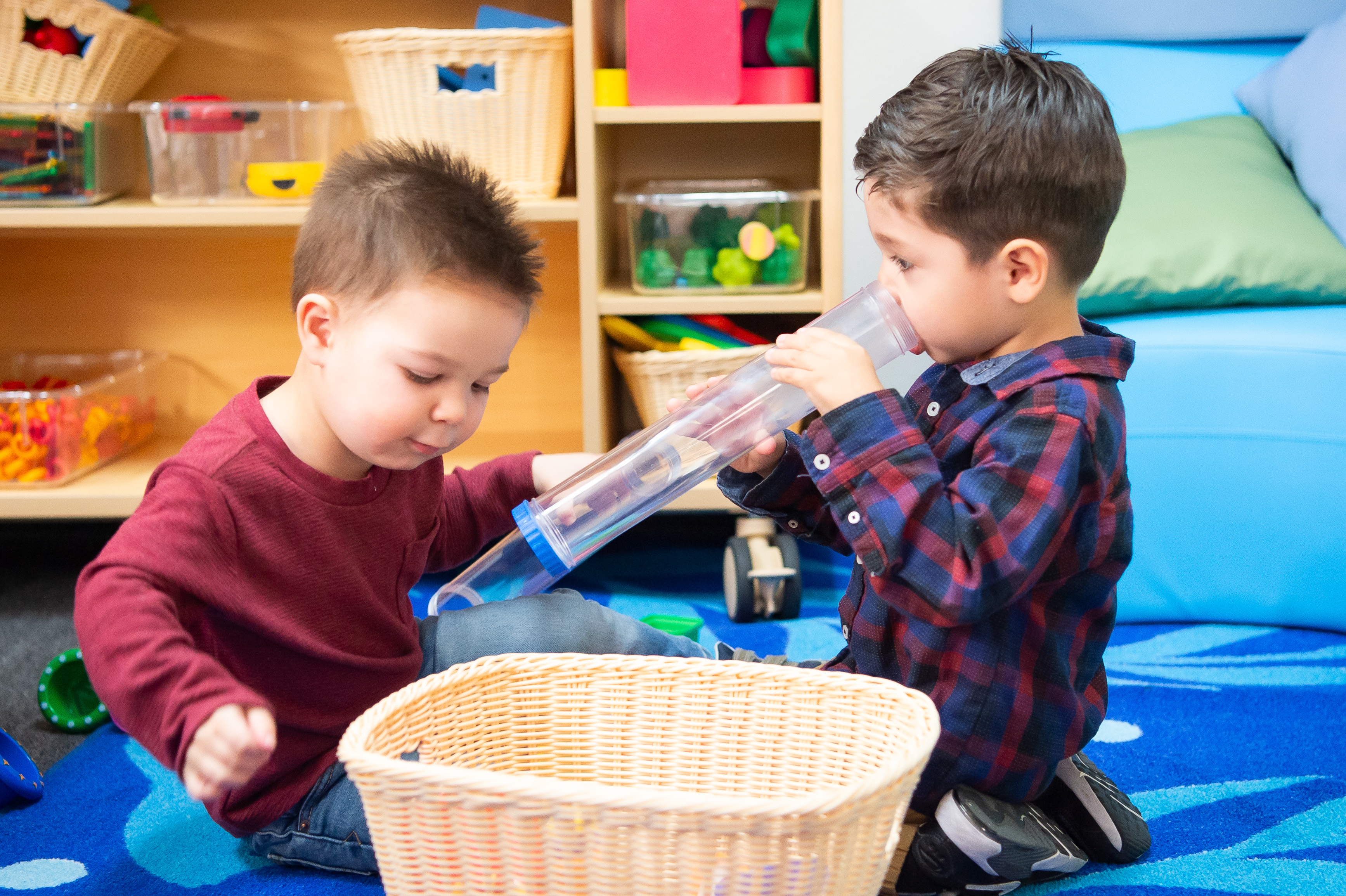 Two young boys, one with a red shirt and one with a blue flannel play with toys in a colorful classroom setting