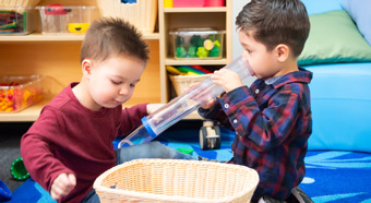 Two young boys, one with a red shirt and one with a blue flannel play with toys in a colorful classroom setting