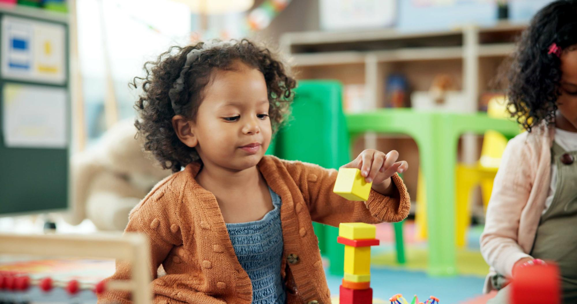 Toddler girl in a classroom setting plays with colorful blocks on the floor, stacking them up, smiling. 