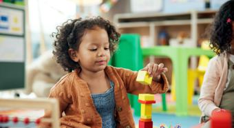 Toddler girl in a classroom setting plays with colorful blocks on the floor, stacking them up, smiling. 