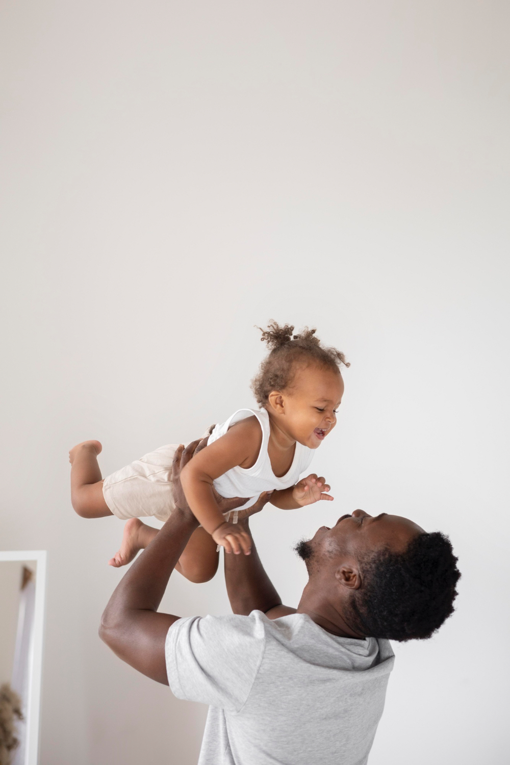 Adult male holds up little toddler girl above his head, both laughing and looking at each other smiling. 