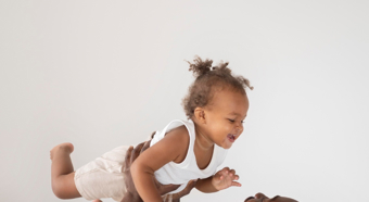 Adult male holds up little toddler girl above his head, both laughing and looking at each other smiling. 
