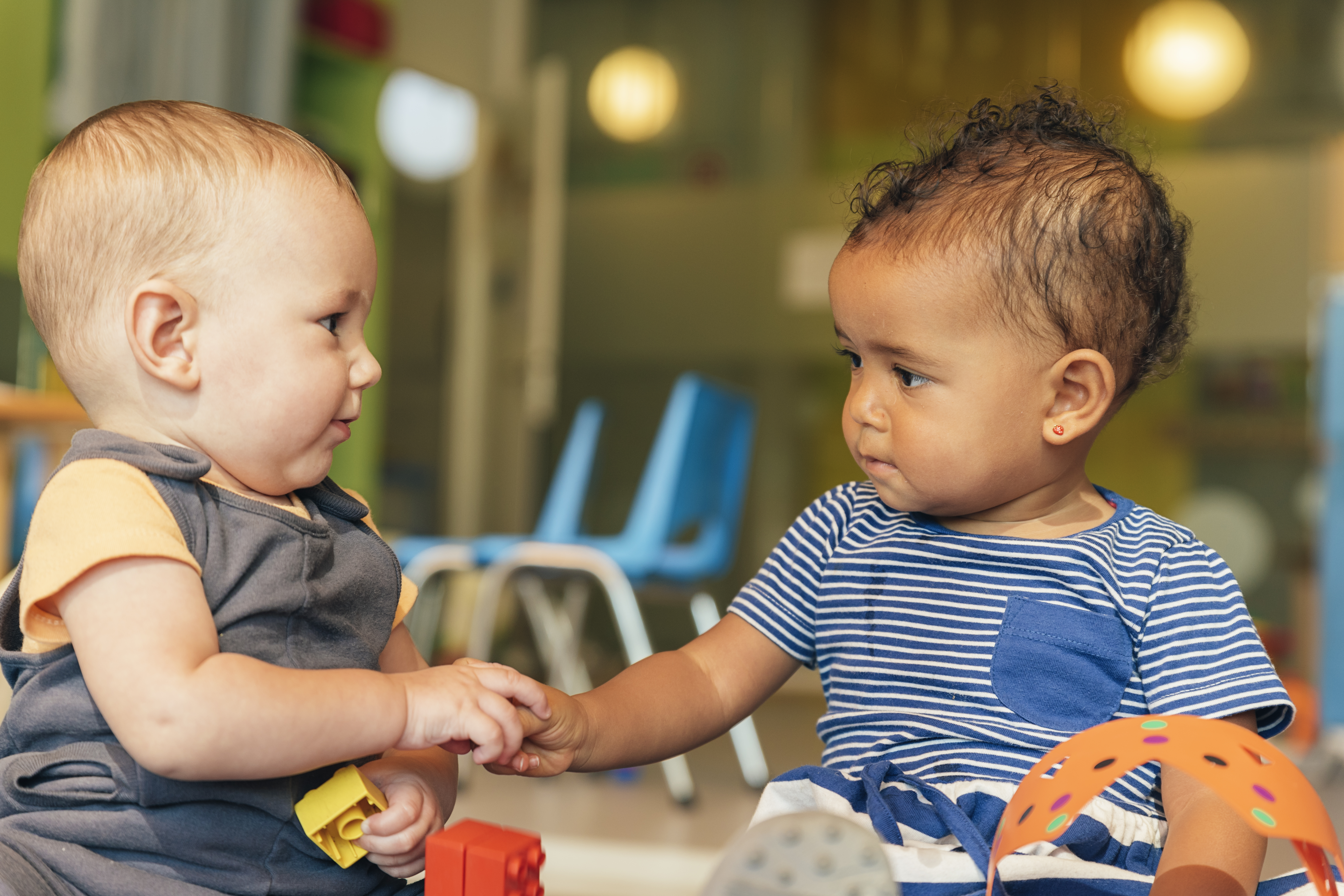 Two toddlers sitting on floor of school setting stare at each other while their hands touch.