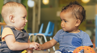 Two toddlers sitting on floor of school setting stare at each other while their hands touch.