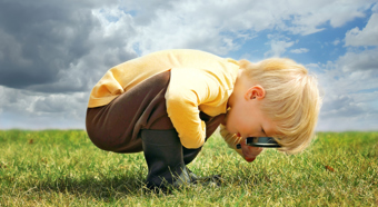 A toddler-aged boy with light skin and blonde hair is bending over looking at the grass with a magnifying glass.