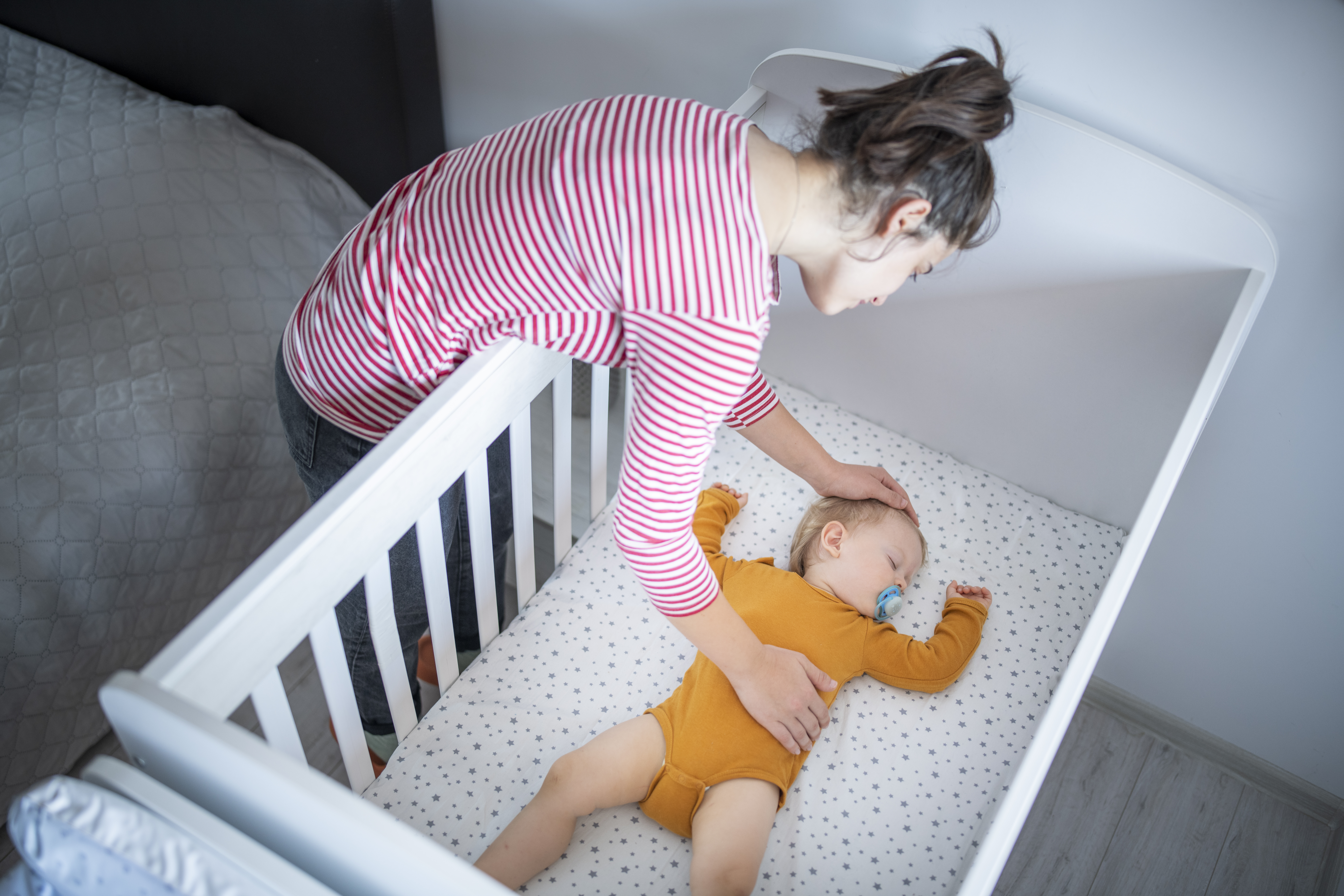 A woman with light skin and brown hair pulled up into a messy bun is leaning over a crib placing her hands gently on a sleeping baby.